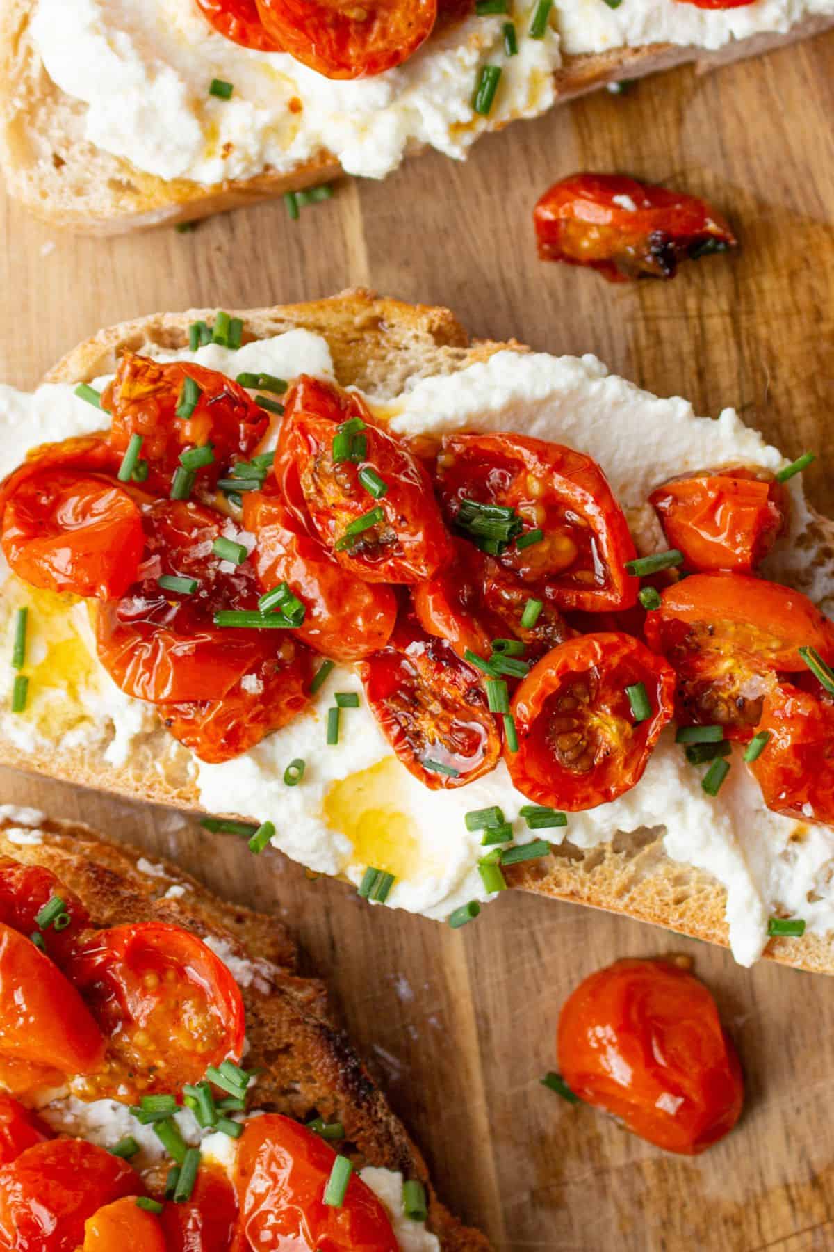 Close up of a slice of 2 slices of toast with cream cheese and baked cherry tomatoes topped with chives.