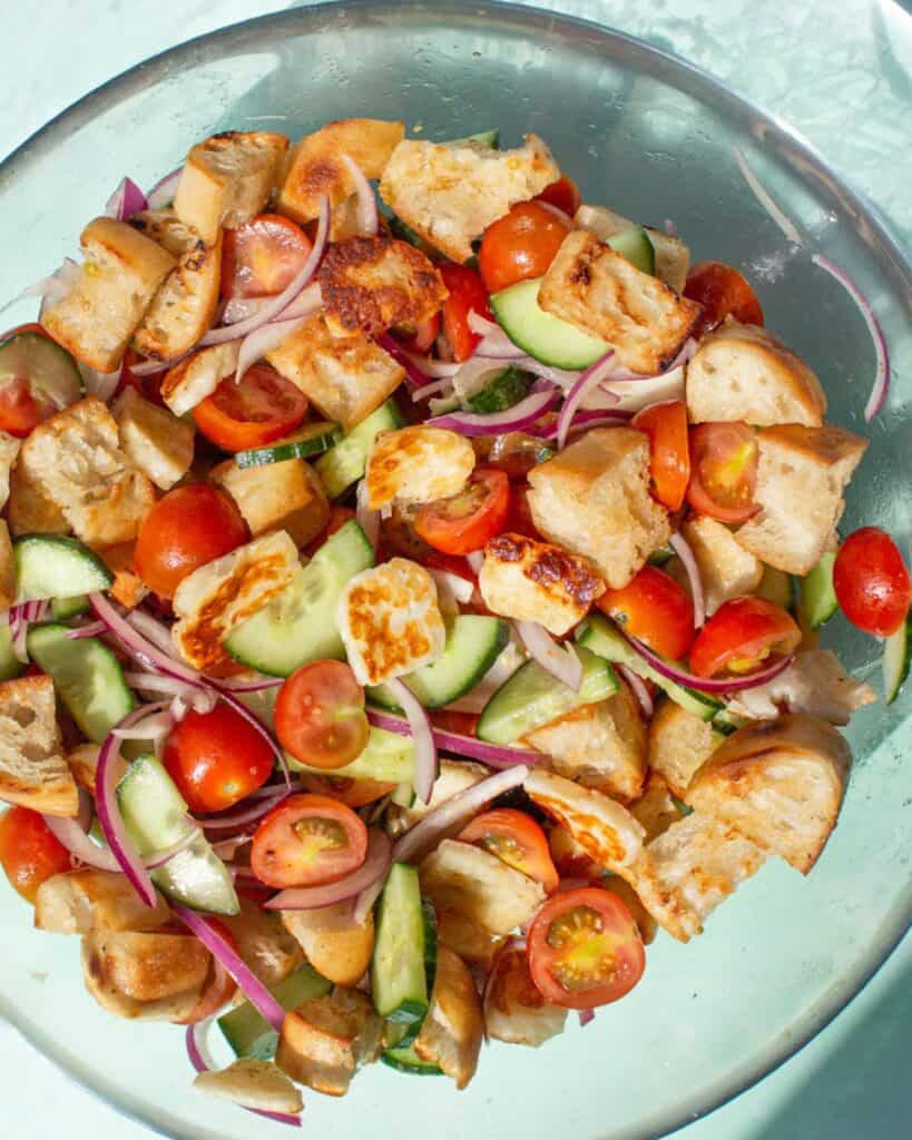 Salad with cherry tomatoes, red onion, bread pieces and fried halloumi in a glass bowl.