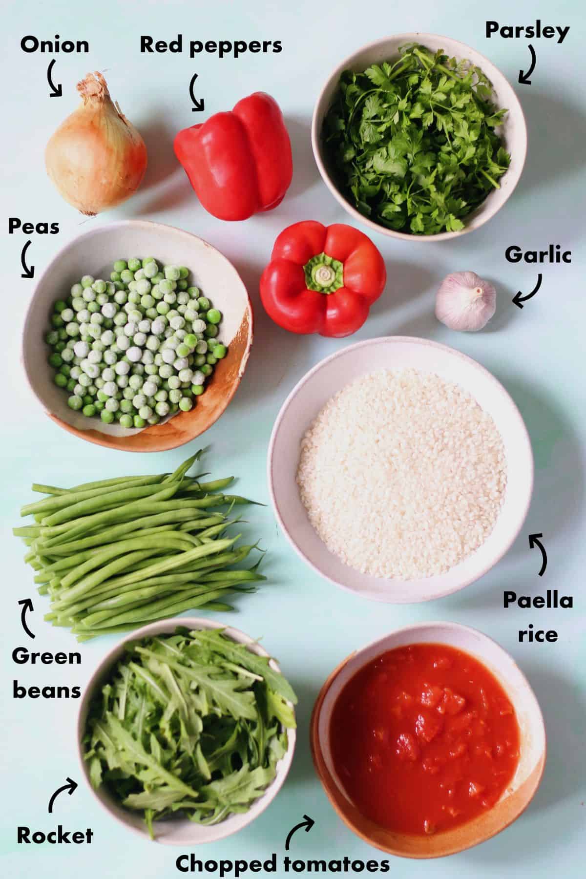 Over head shot of ingredients to make paella laid out on a pale blue background and labelled.