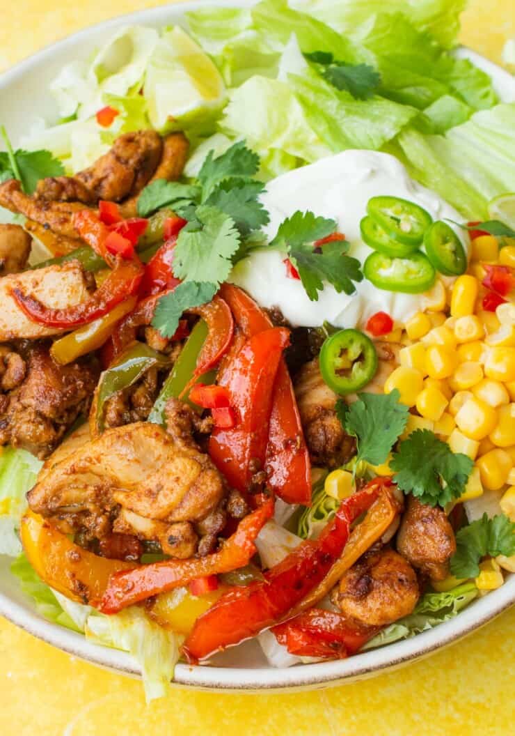 Close up of Chicken Fajita Salad with lettuce, sweetcorn, sour cream, chicken fajita mix with peppers and green chilli slices in a white bowl.