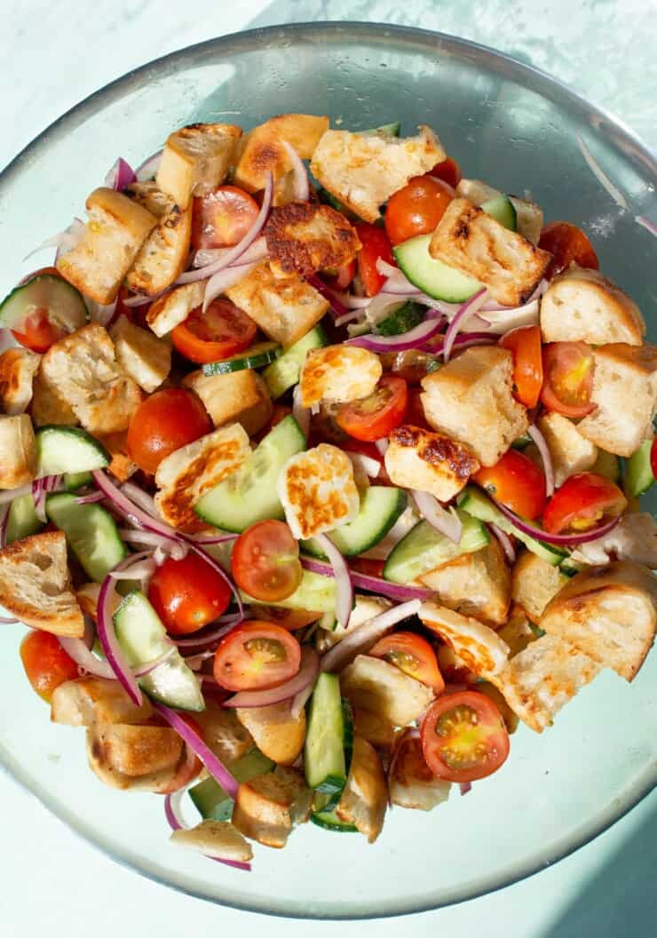 Halloumi Panzanella salad with cherry tomatoes, cucumber, red onion, browned halloumi and ciabatta bread cubes in a large glass bowl.