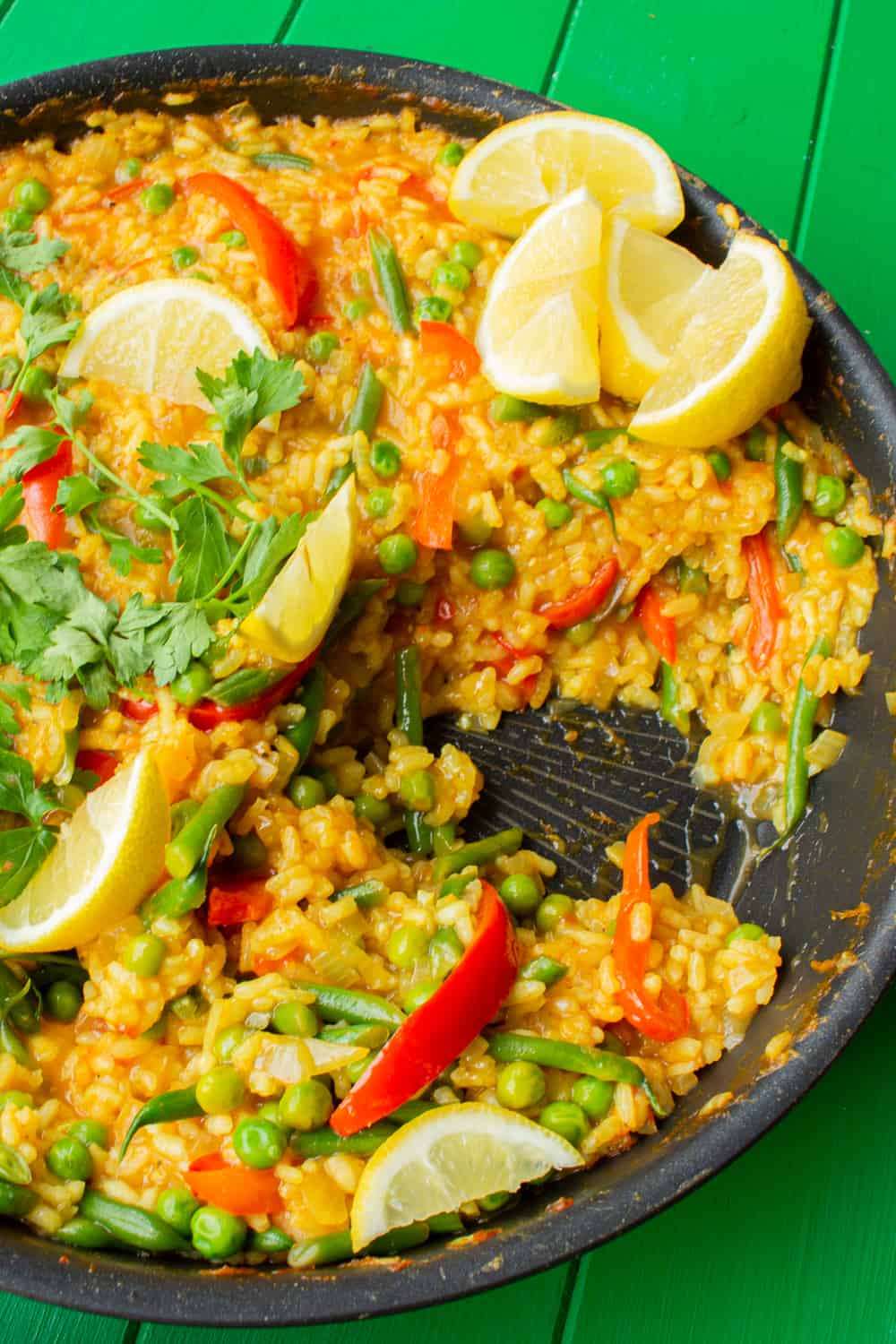 Vegetable paella with peppers, peas and beans and topped with lemon wedges and fresh parsley and a portion already served out of pan.