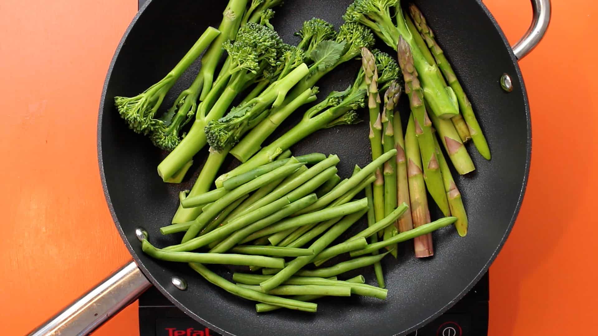 Broccoli stems and asparagus in large frying pan on a stove on an orange background.