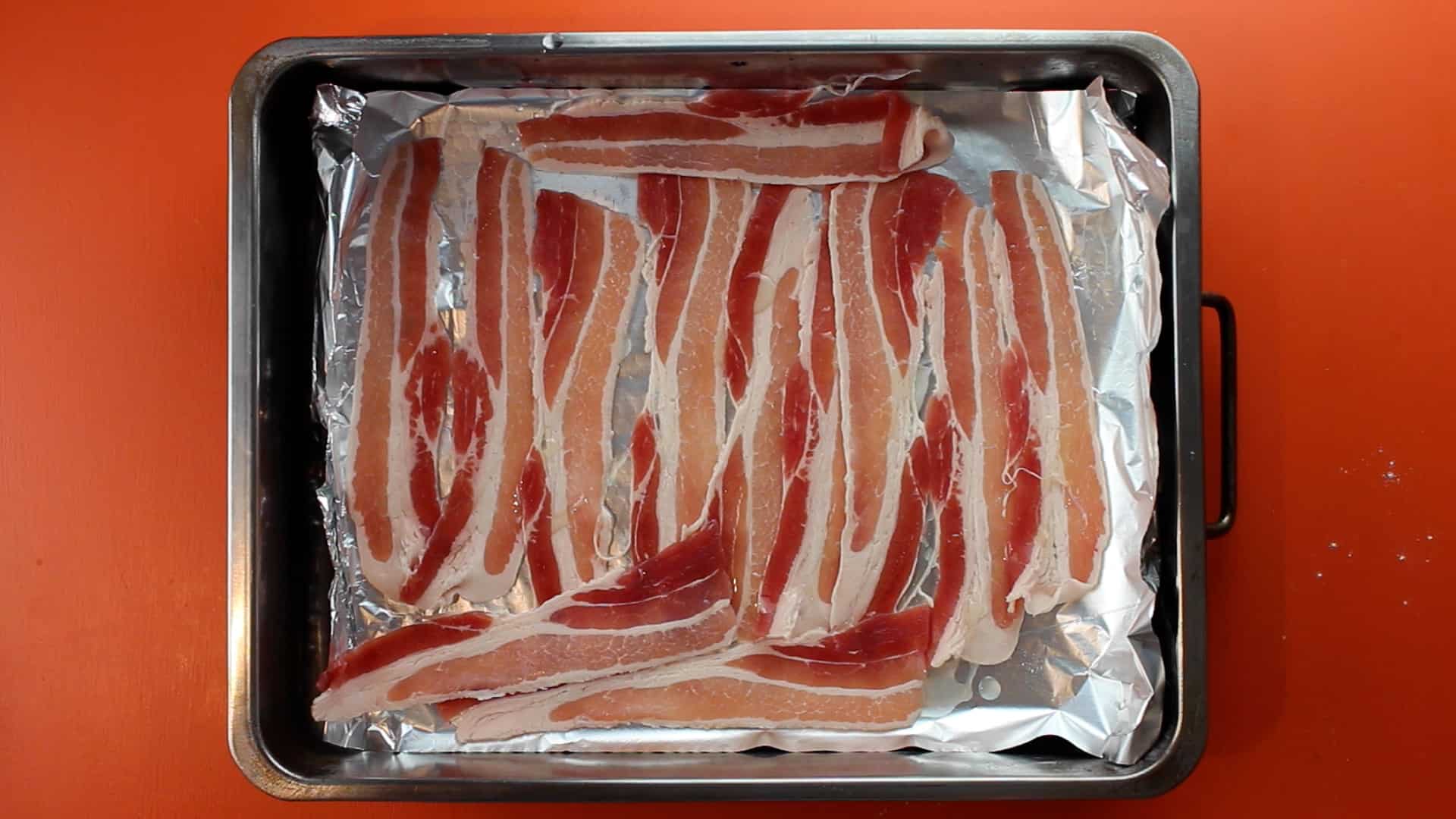 Rashers of bacon on a foil lined baking tray on an orange background.