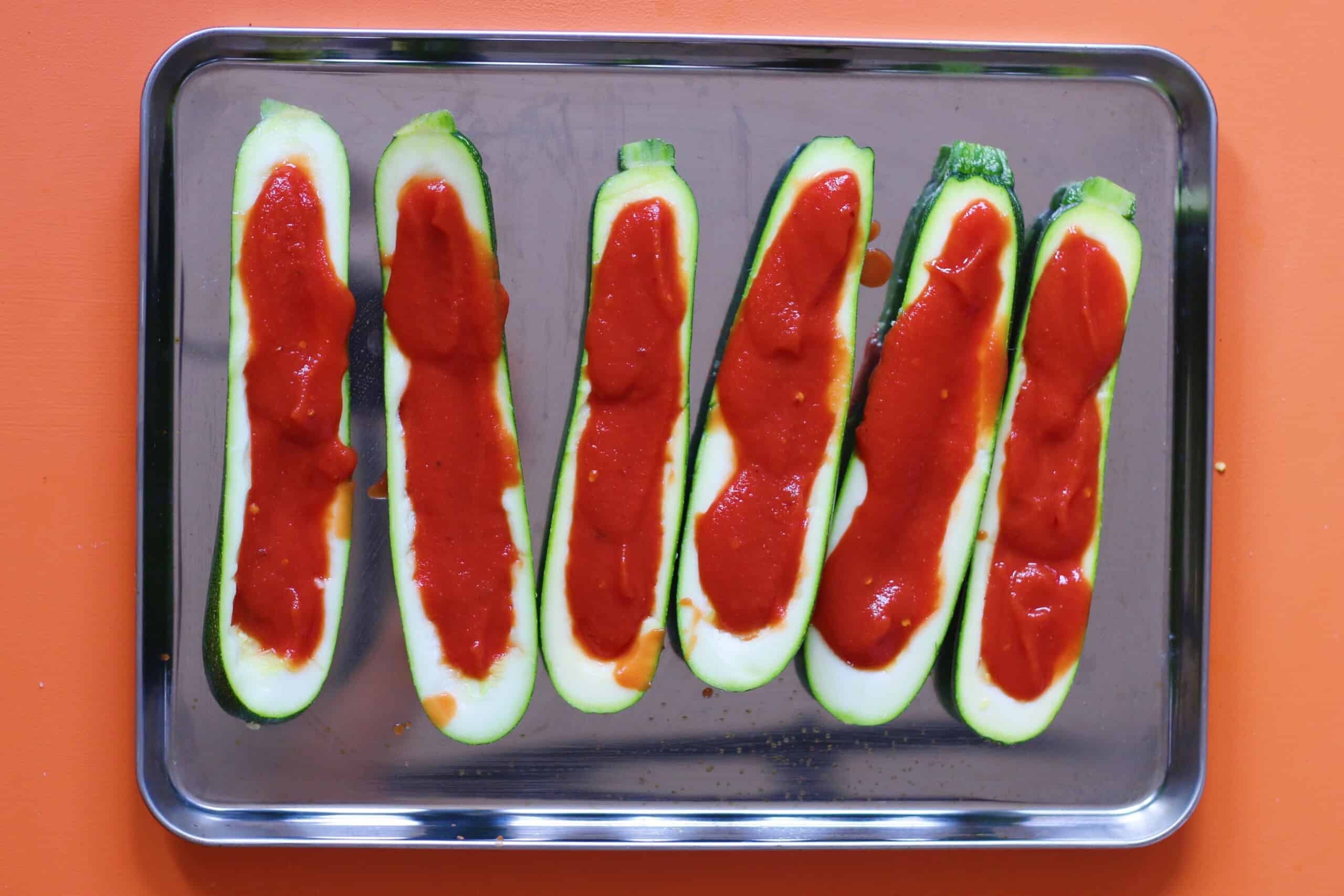 6 courgette boats filling with passata on a stainless steel baking tray on an orange background.
