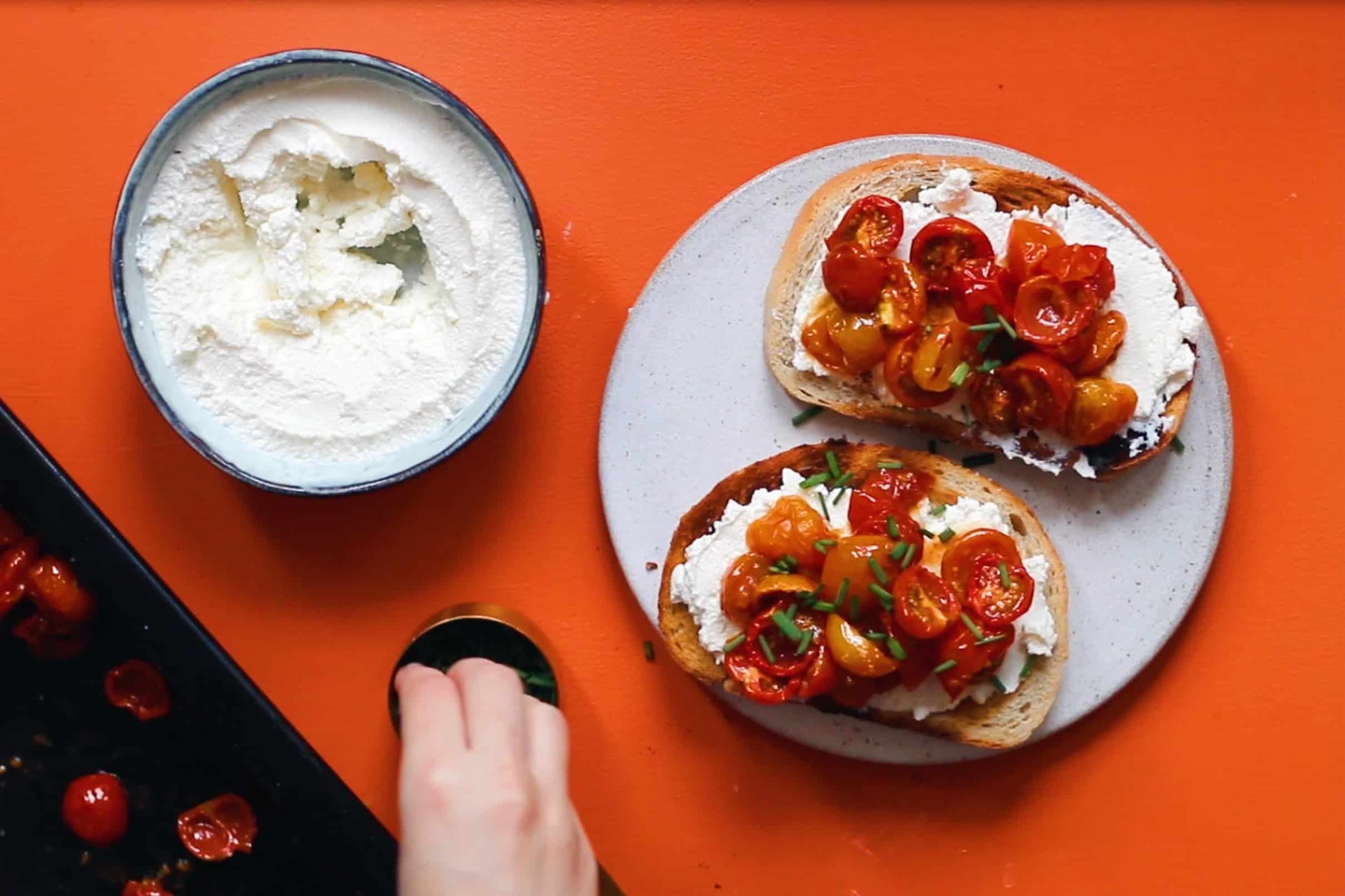 Roasted cherry tomatoes and ricotta served on 2 pieces of toasted sourdough on a plate next to chives and a bowl of ricotta. 