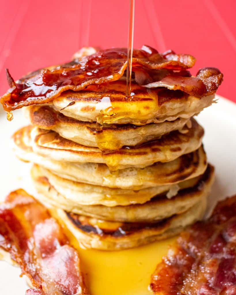 American Style Pancakes stacked on top of each other, topped with crispy bacon and maple syrup poured on pancakes and bacon pieces on plate.