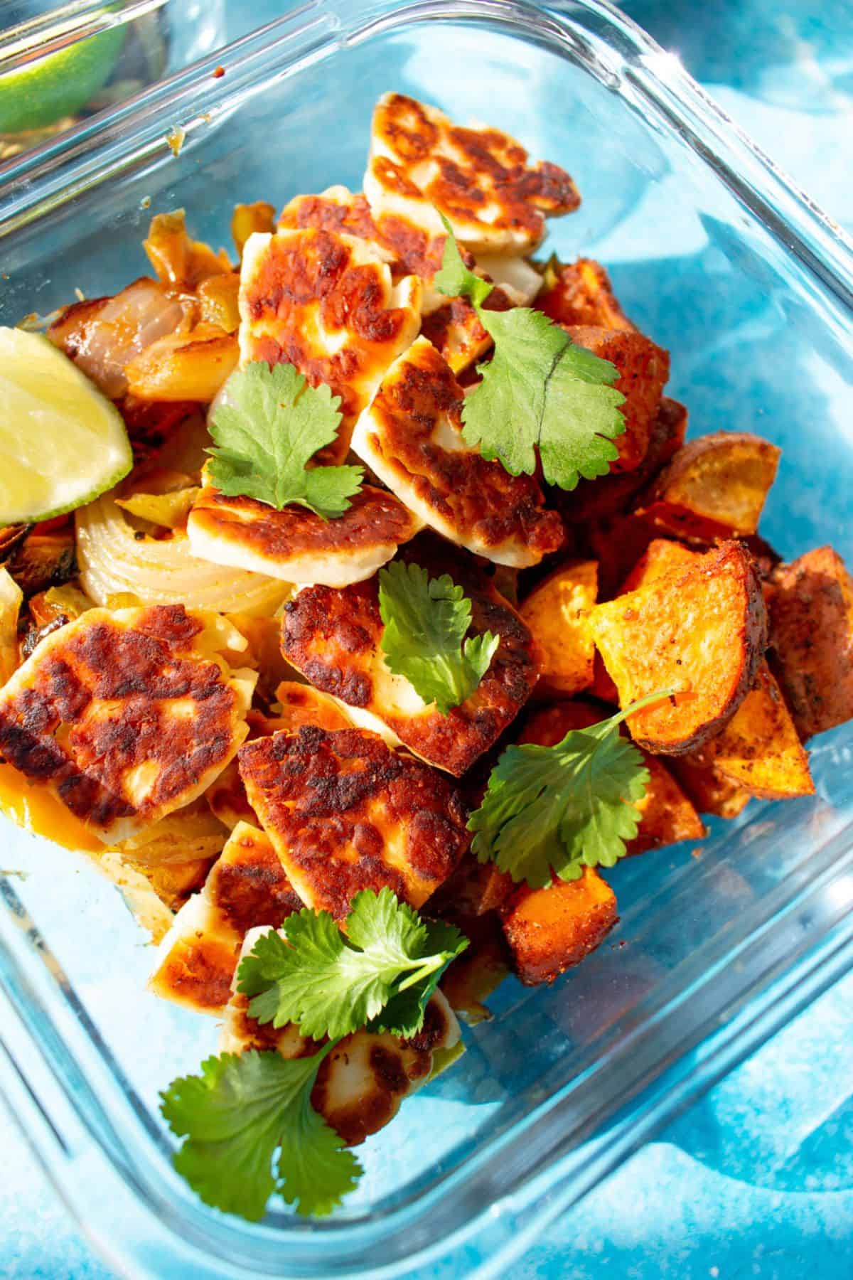 Close up shot of a square glass meal prep container filled with sweet potato cubes, peppers, topped with golden browned halloumi, coriander and a lime wedge.