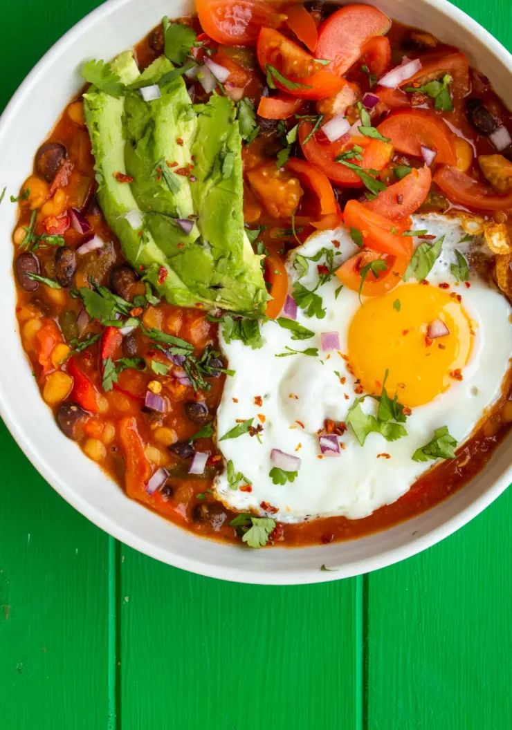 Huevos Rancheros inspired Baked Beans in a white bowl with avocado slices, tomatoes, a fried egg on the mixed beans on a green background.