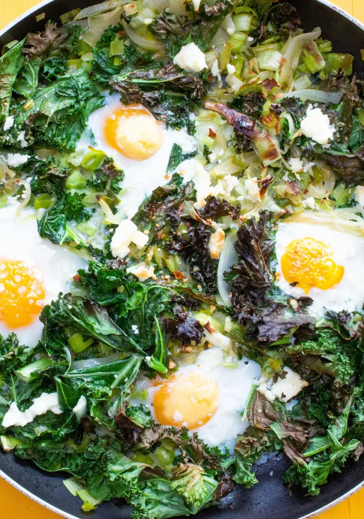 Green Shakshuka with leeks, kale and 4 eggs in a large pan on a yellow background.