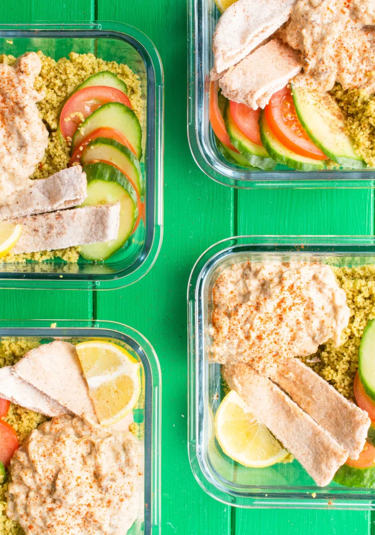 4 square glass meal prep containers with Baba ganoush; humous, cous cous, pitta bread and tomatoes and cucumber.