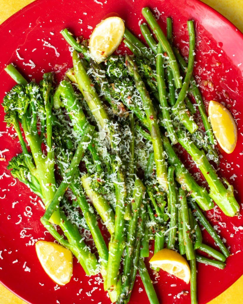 Tenderstem Broccoli with Garlic Butter and asparagus topped with grated parmesan and lemon wedges on a large red plate on a blue background.
