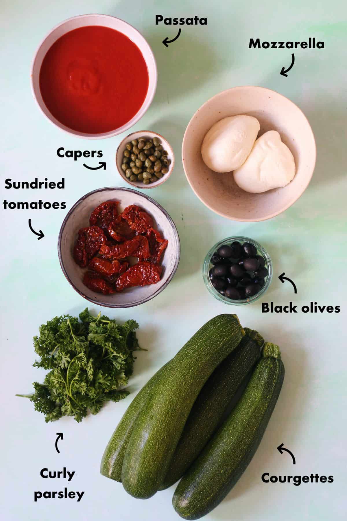 Ingredients to make Courgette Pizzas with sundried tomatoes laid out on a pale blue background and labelled.
