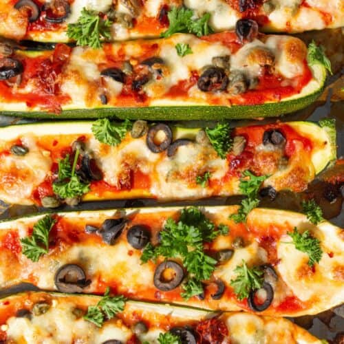 CUrgette halves roasted with tomatomsauce and toppings of olives and capers with parsley on a baking tray.