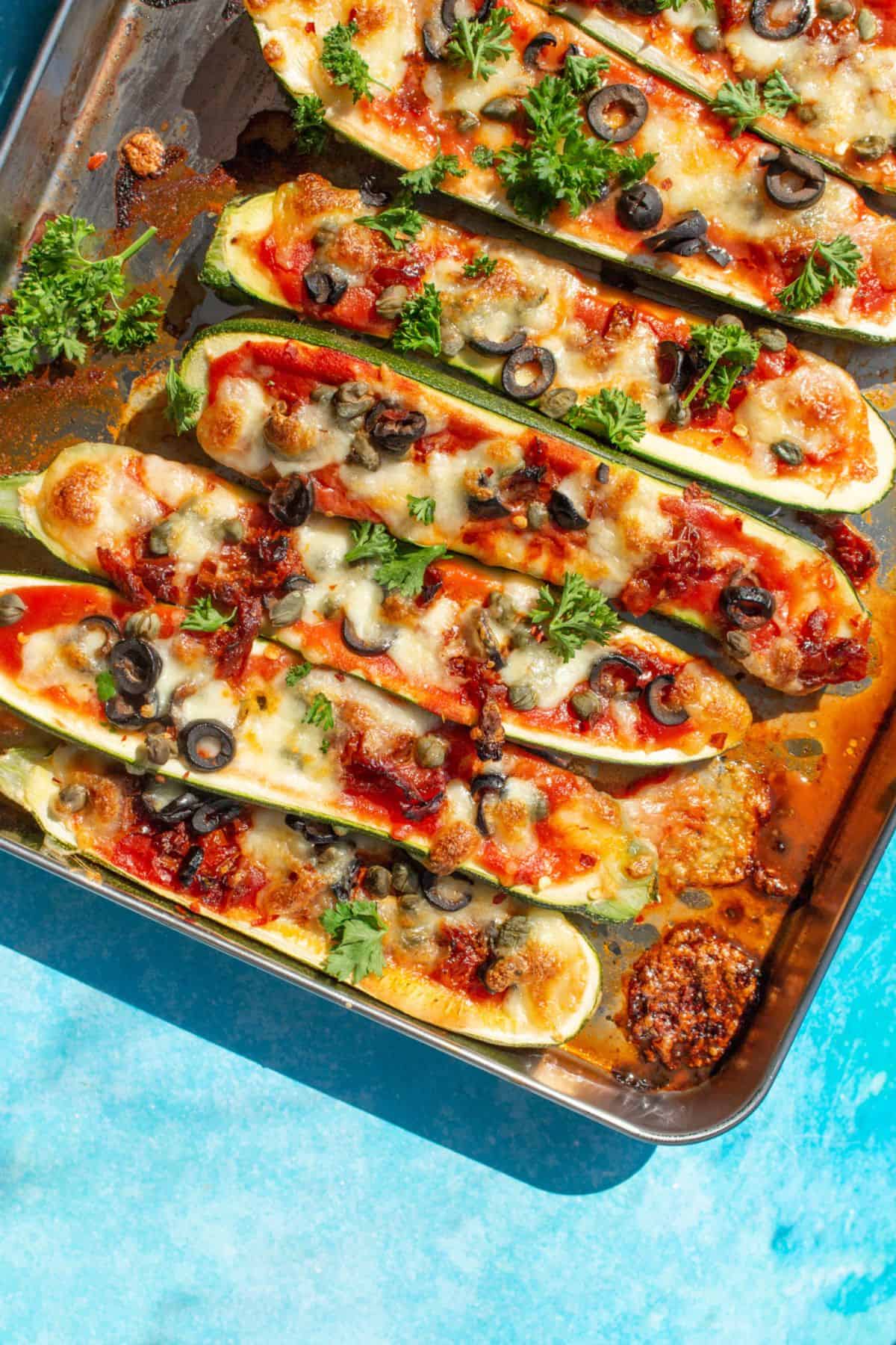 Courgette halves on a baking tray with golden brown cheese topping , sliced black olives and fresh parsley on a blue background.