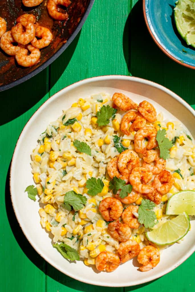 A bowl with Sweetcorn Orzo with Paprika Prawns with coriander and lime wedges with partial view of pan with prawns and bowl of lime wedges.