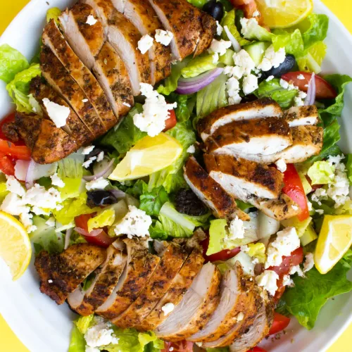 Greek chicken salad in a bowl with slices of chicken, lettuce, tomato, red onion and lemon wedges.