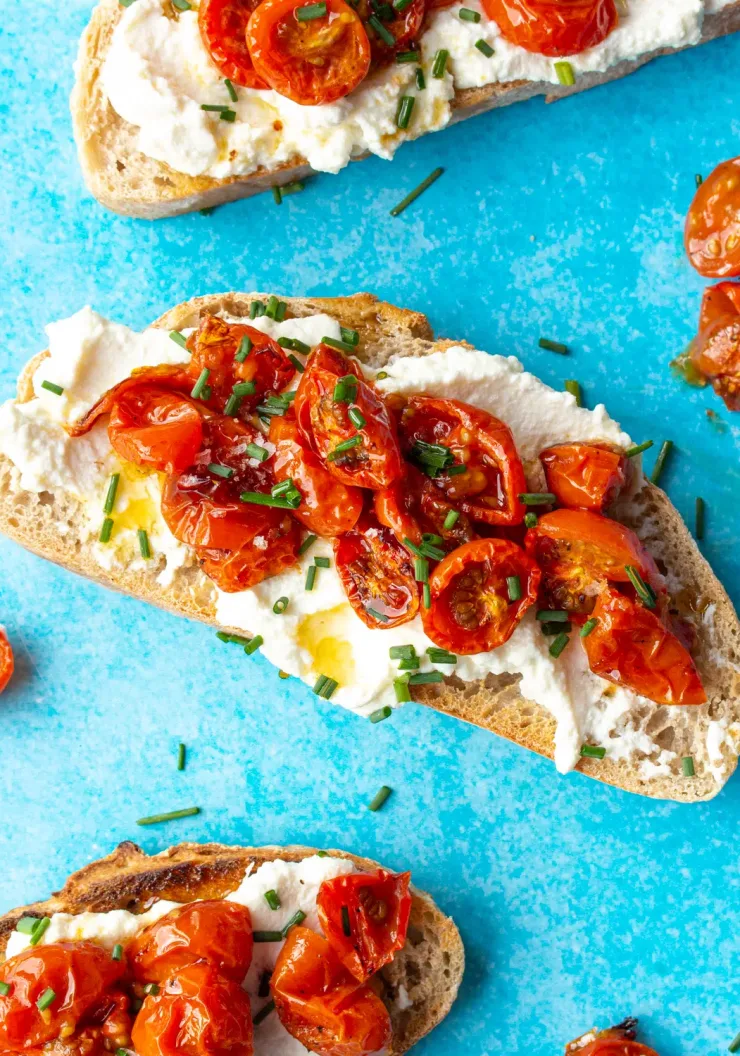 Tomato & Ricotta toast with 3 pieces of sourdough with ricotta, cherry tomatoes and chives on a turquoise background.