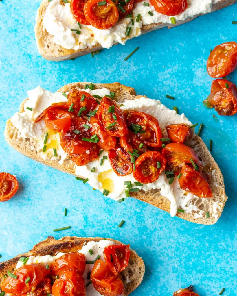 Tomato & Ricotta toast with 3 pieces of sourdough with ricotta, cherry tomatoes and chives on a turquoise background.