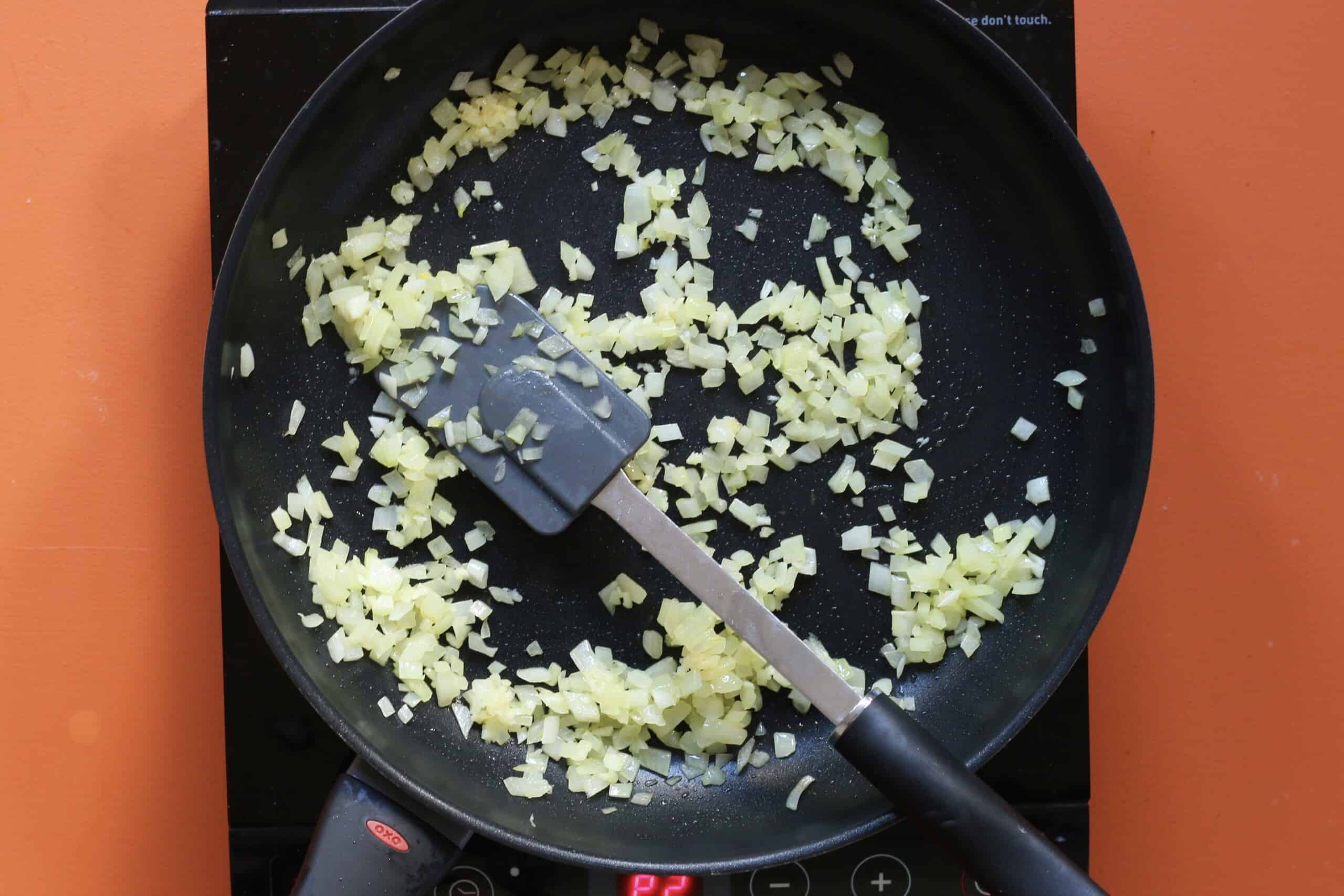 Diced onion sautéing in a frying pan  and stirred with a spatula on a stove on an orange background.