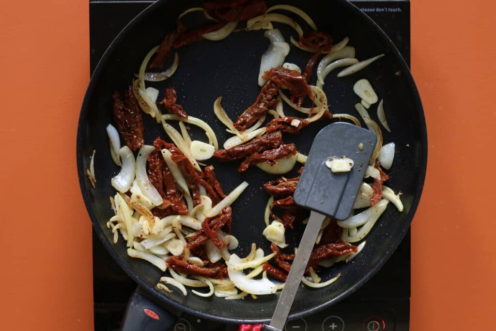 Sundreid tomatoes and sliced onions frying and stirred with a spatula in a pan on a stove on an orange background.