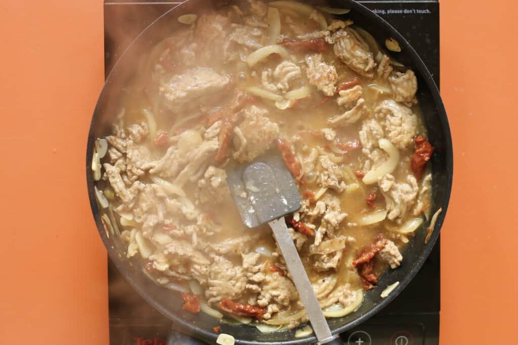 Pork mince, sun-dried tomatoes, onions and stock mixed with a spatula in a pan on a stove on an orange background.