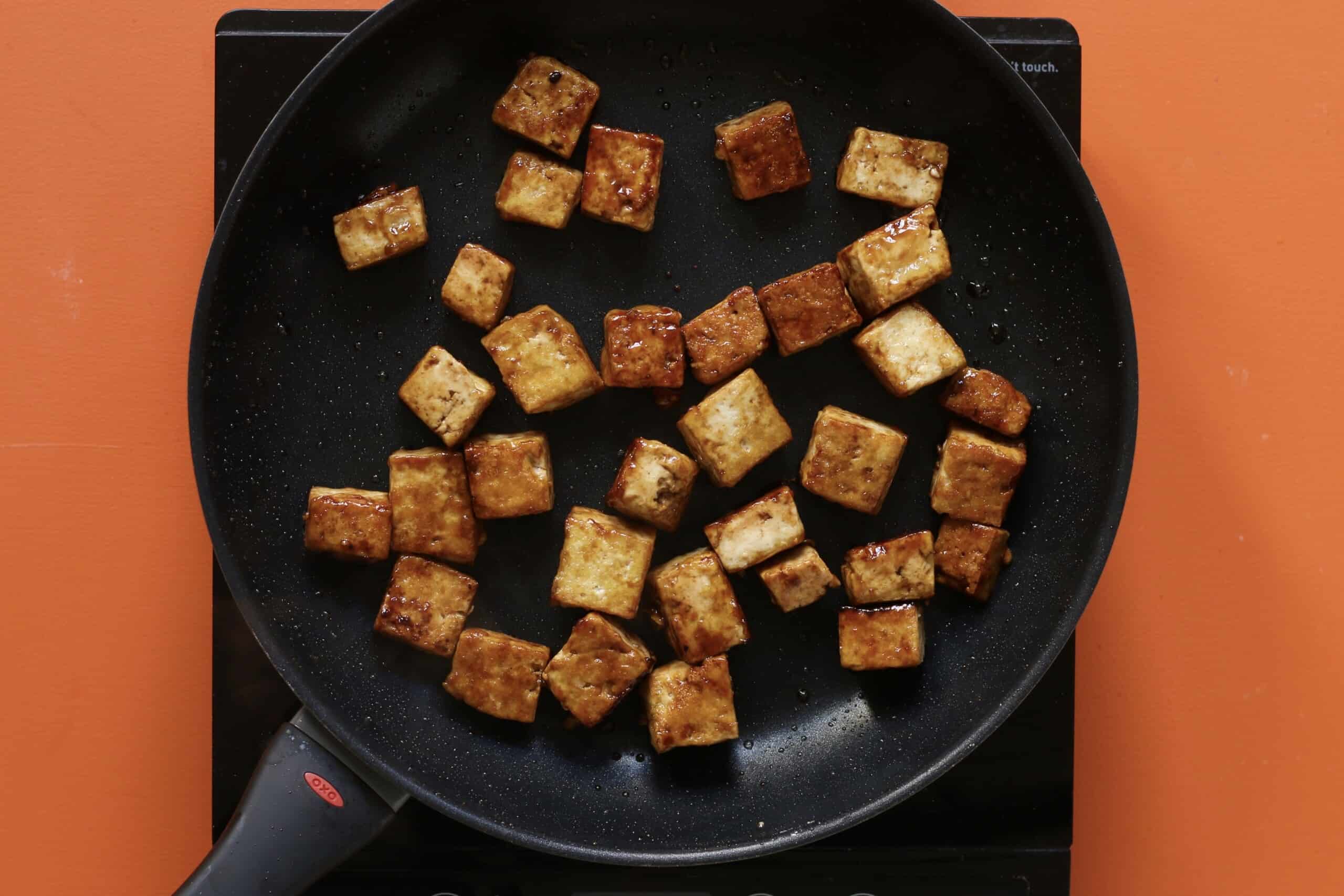 Golden browned tofu cubes in frying pan on stove on an orange background.