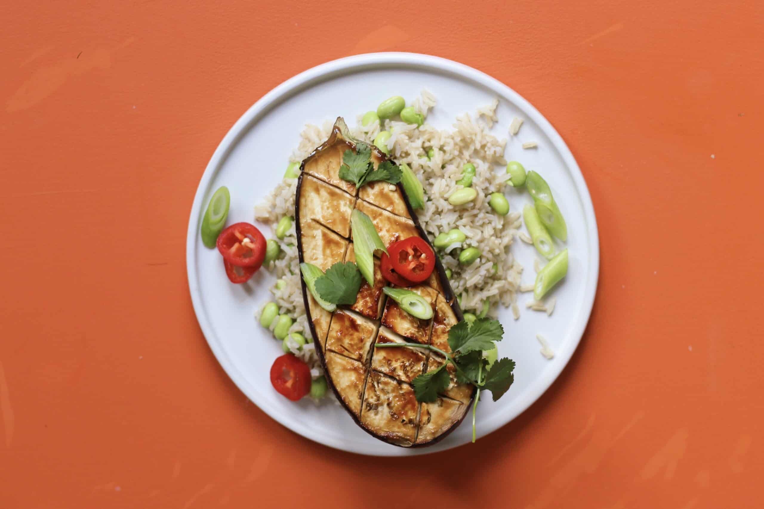 Half a roasted Miso aubergine on a bed of rice with edamame beans and spring onions on a plate on an orange background.
