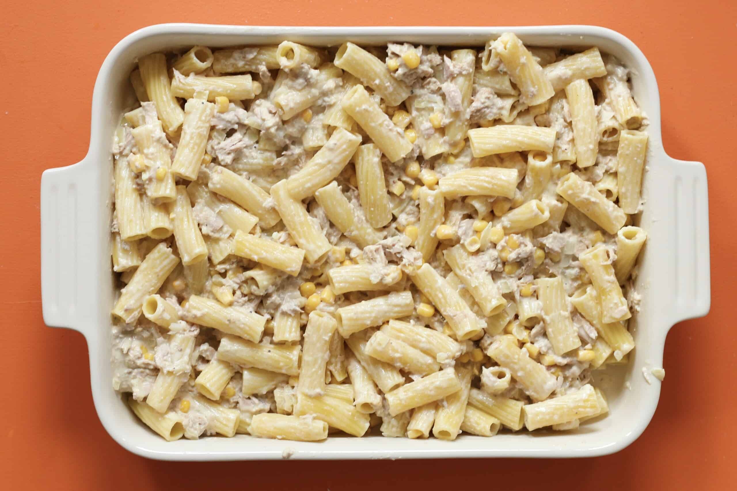 Rigatoni, sweetcorn and cheesy sauce mixed together in a white rectangular baking dish on a stove on an orange background.