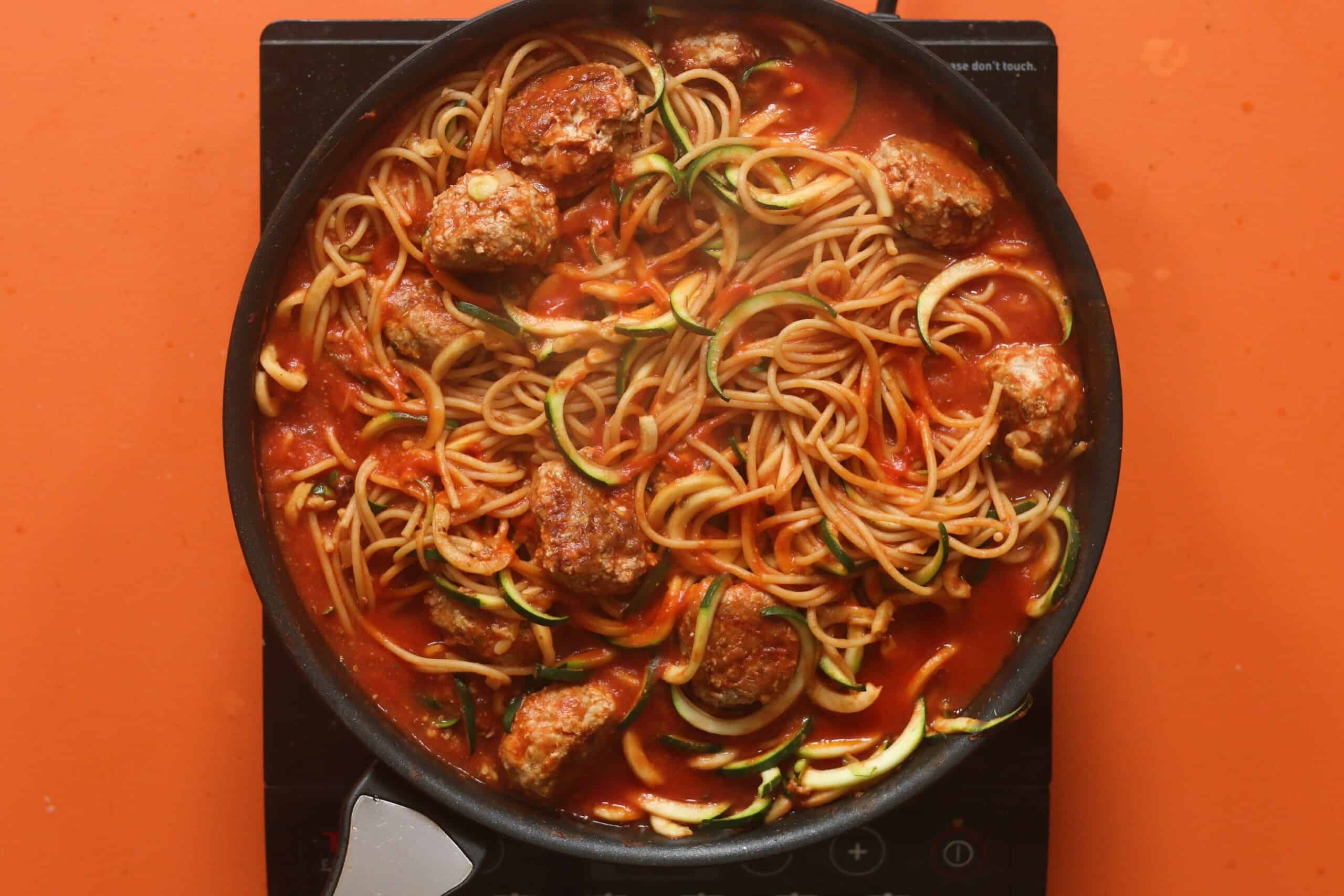 Meatballs added to the spaghetti and spiralised courgettes in tomato sauce in the pan on the stove on an orange background.