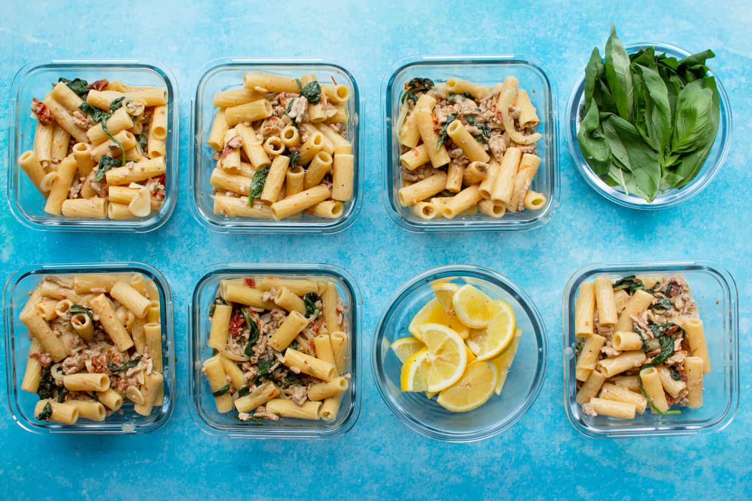 6 square glass meal prep containers with Tuscan inspired rigatoni with pork mince and 2 round glass bowls with lemon wedges and fresh basil.