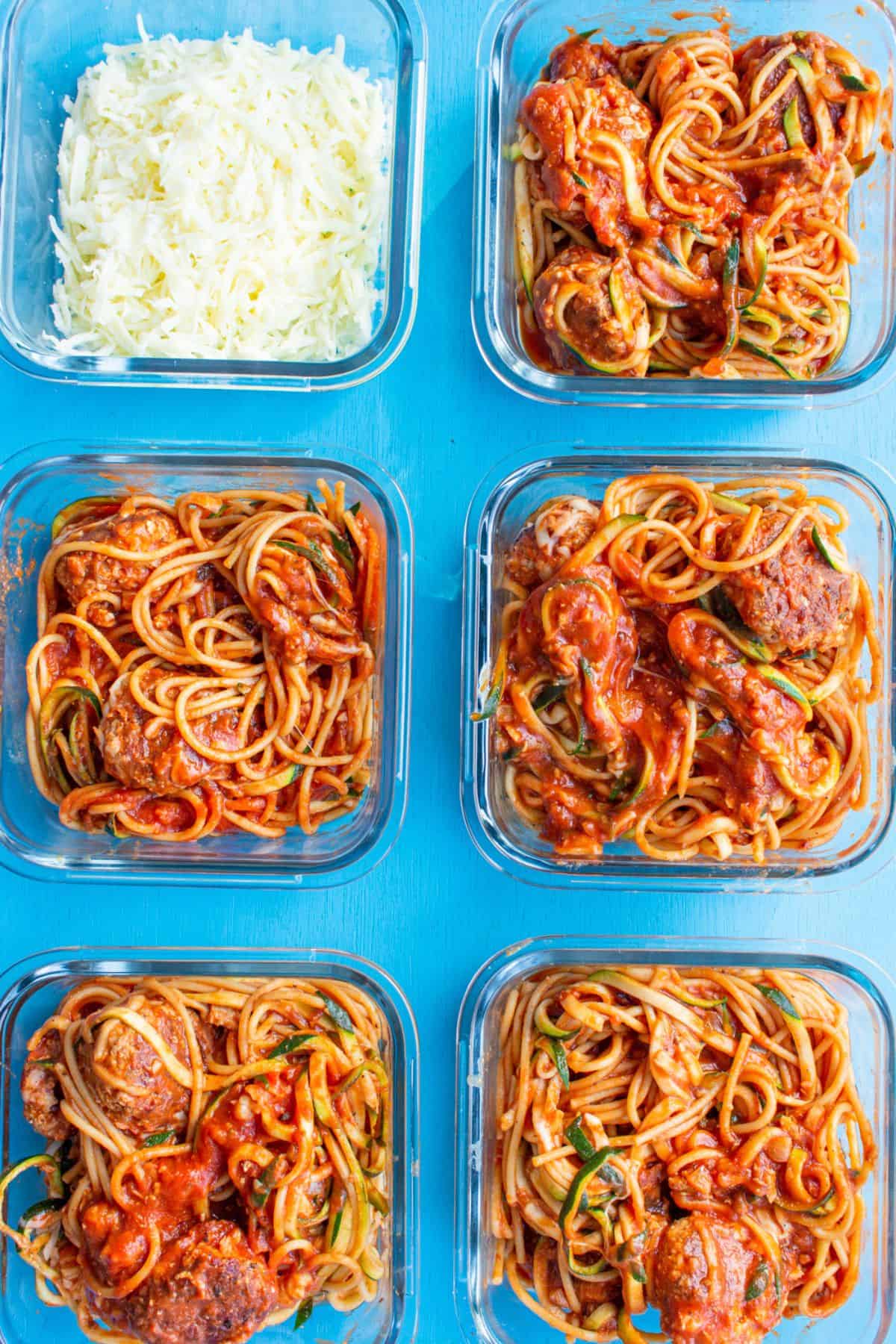 6 square glass meal prep containers, one with grated cheese and the other with meatball marinara and spaghetti in containers.
