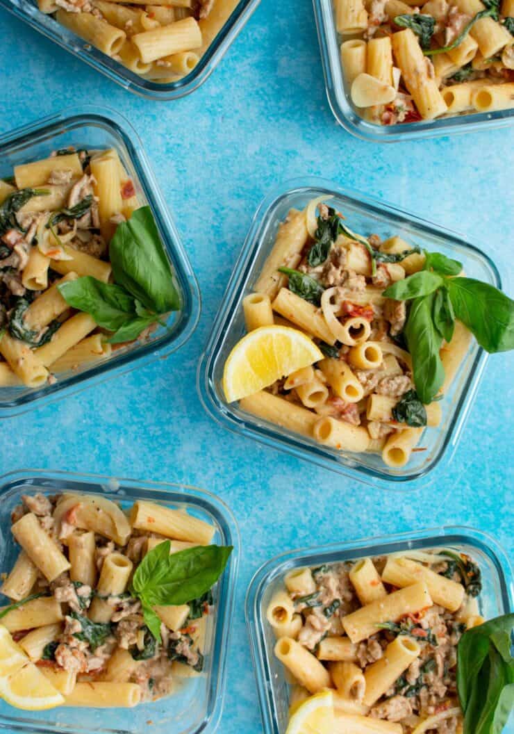 6 square glass meal prep containers filled with creamy rigatoni with pork mince garnished with fresh basil and lemon wedges.