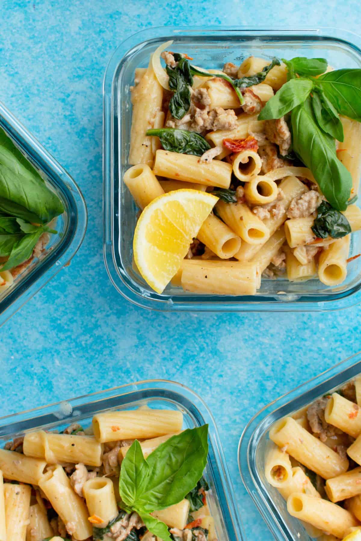 Glass meal prep containers filled with rigatoni with pork mince garnished with fresh basil and lemon wedges.