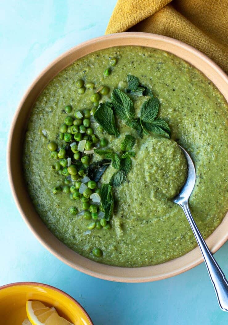 Pea soup with fresh mint scatters over the top and whole peas in a bowl with a spoon on a mustard cloth.