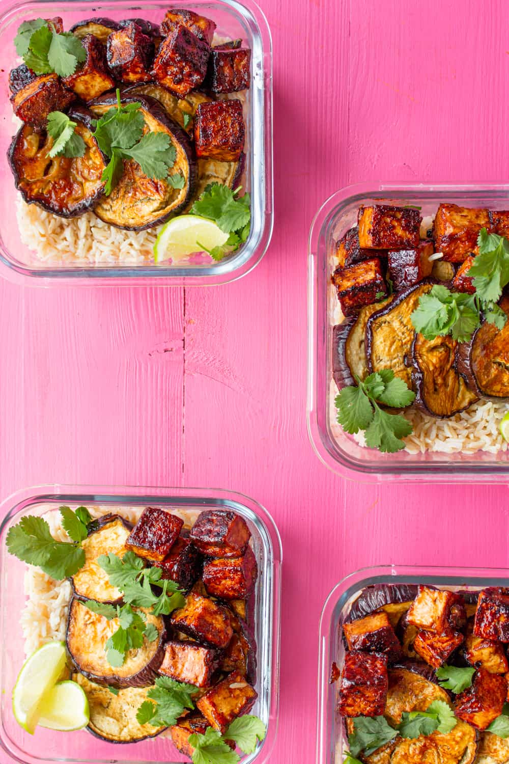 Aubergine and crispy soy tofu & rice in 4 meal prep containers topped with coriander and lime wedges on a pink background.