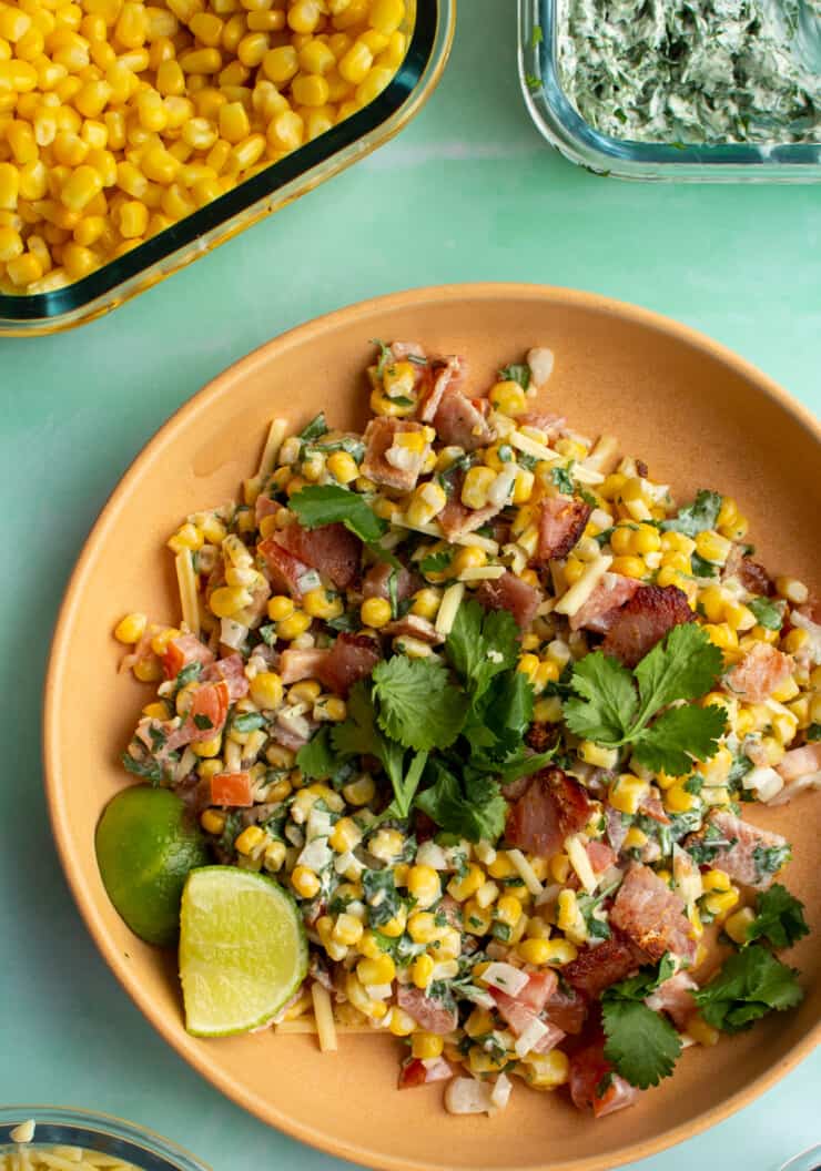 Elote Inspired Bacon Salad in a bowl with sweet corn, coriander and lime wedges next to a dish of sweet corn and one with coriander cream.