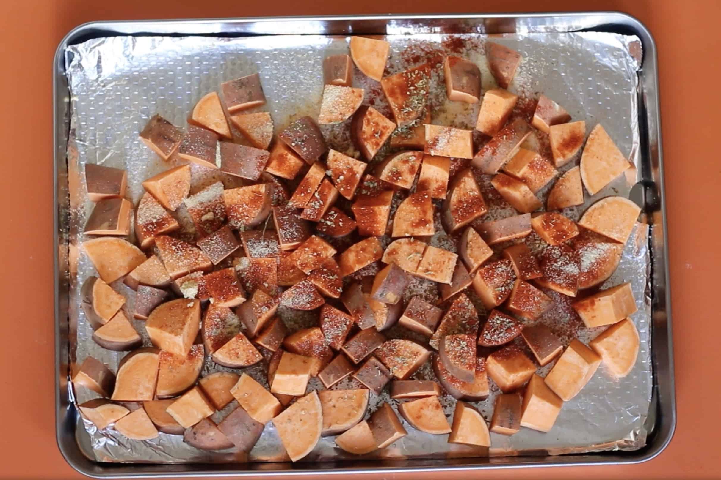 Sweet potatoes cubes with spices added on a foil lined stainless steel baking tray on an orange background.
