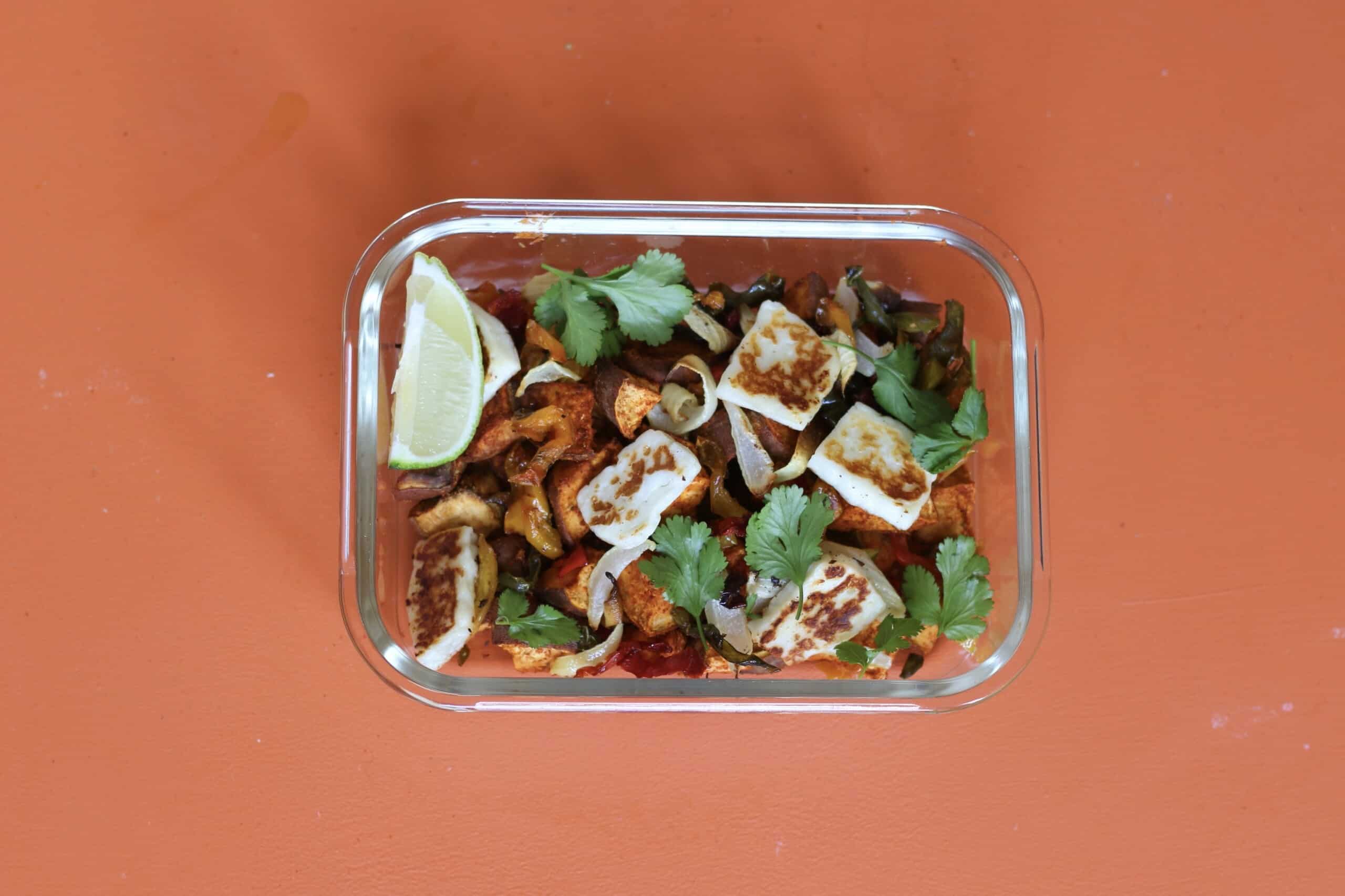 Tex Mex sweet potatoes and halloumi with coriander and a lime wedge in a glass rectangular meal prep container on an orange background.