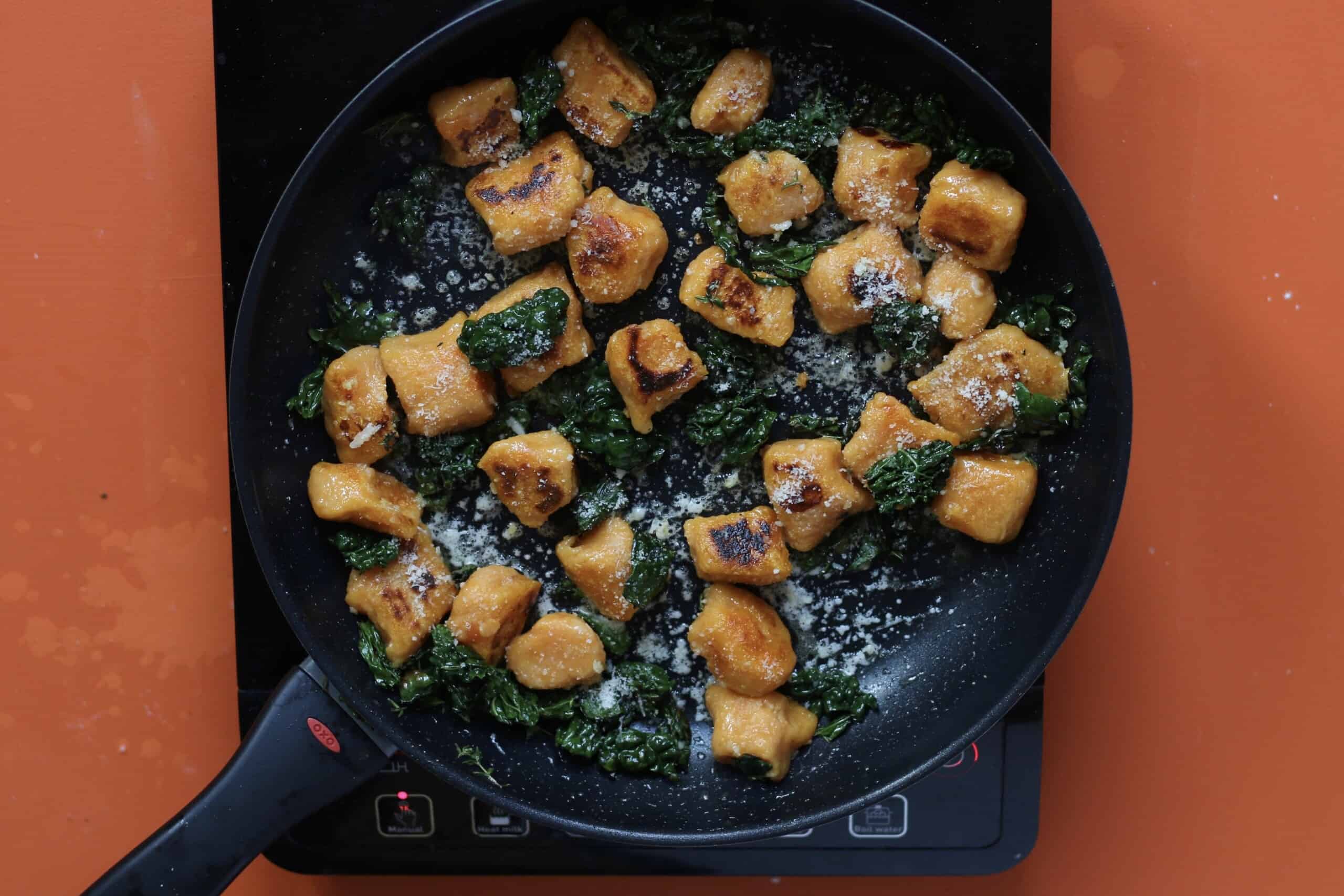 Seet potato gnocchi with cavolo nero browning in a frying pan on a stove on an orange background.