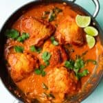 Chicken thigh curry in a pan with sauce and topped with coriander and lime wedges.