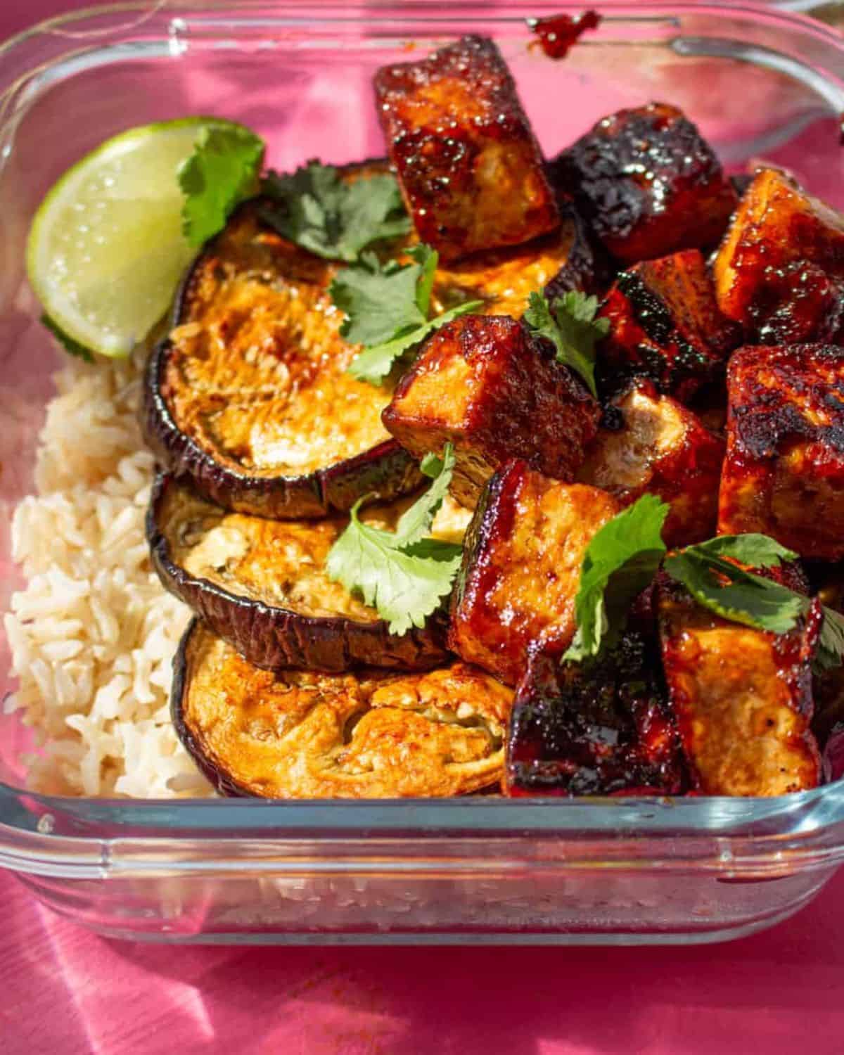 A square, glass meal prep container filled with rice, roasted aubergine rounds, cripsy tofu and a wedge of lemo and topped with coriander..