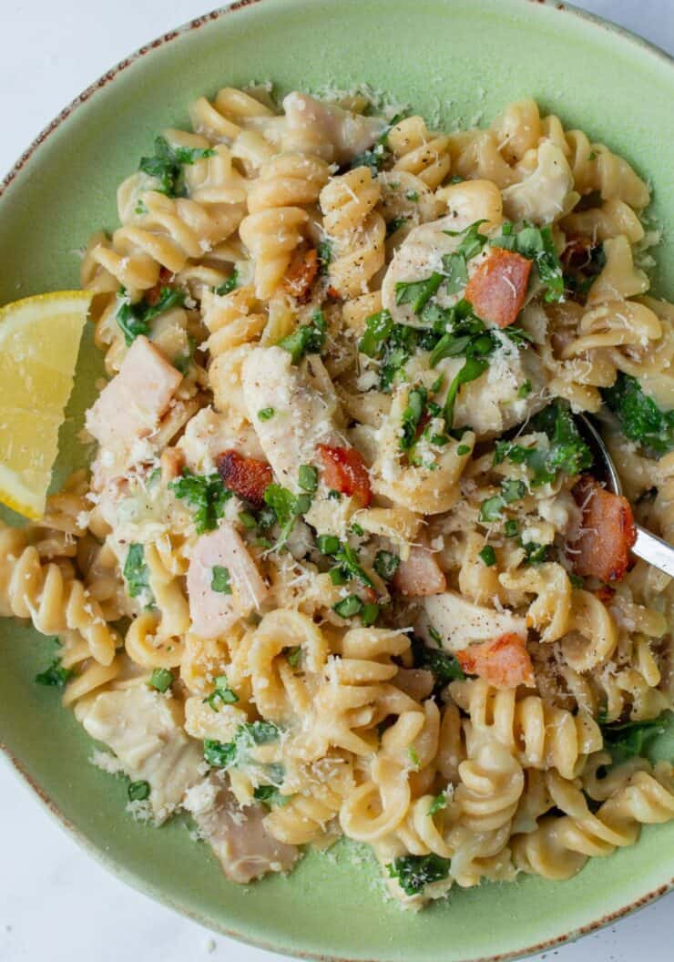 A green bowl with pasta with chicken and bacon in a creamy sauce and a lemon wedge.