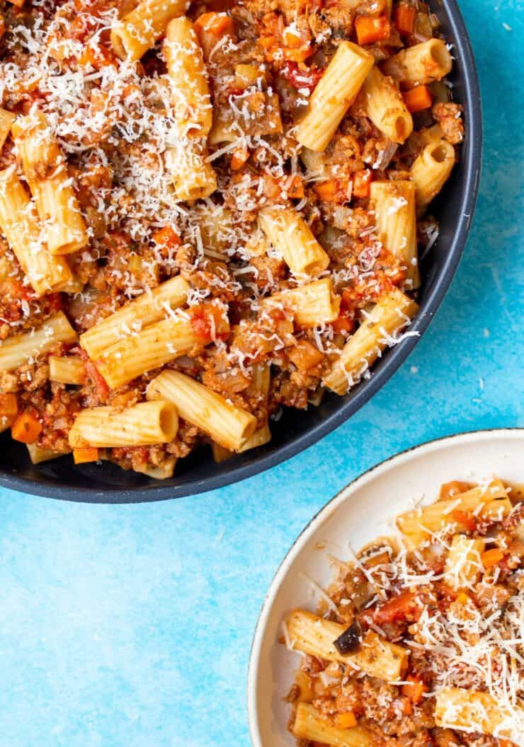 A pan with bolognese with rigatoni pasta and parmesan shavings with a bowl of bolognese pasta.