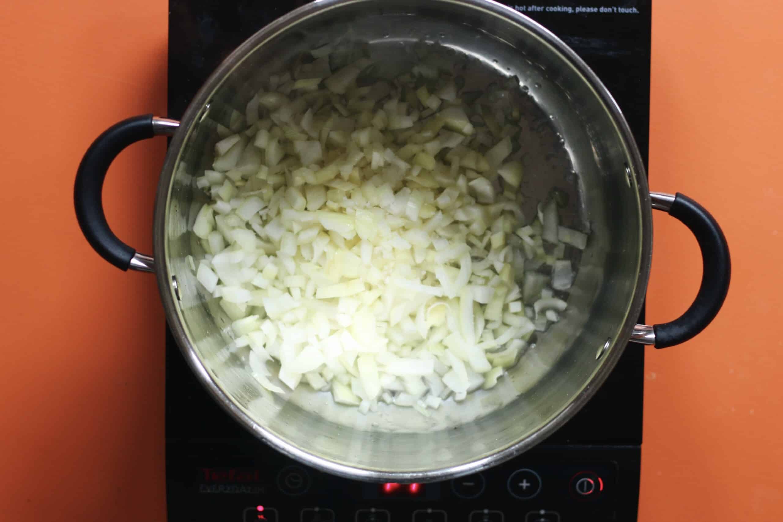 Diced onions frying in a saucepan with 2 handles on a stove on an orange background.