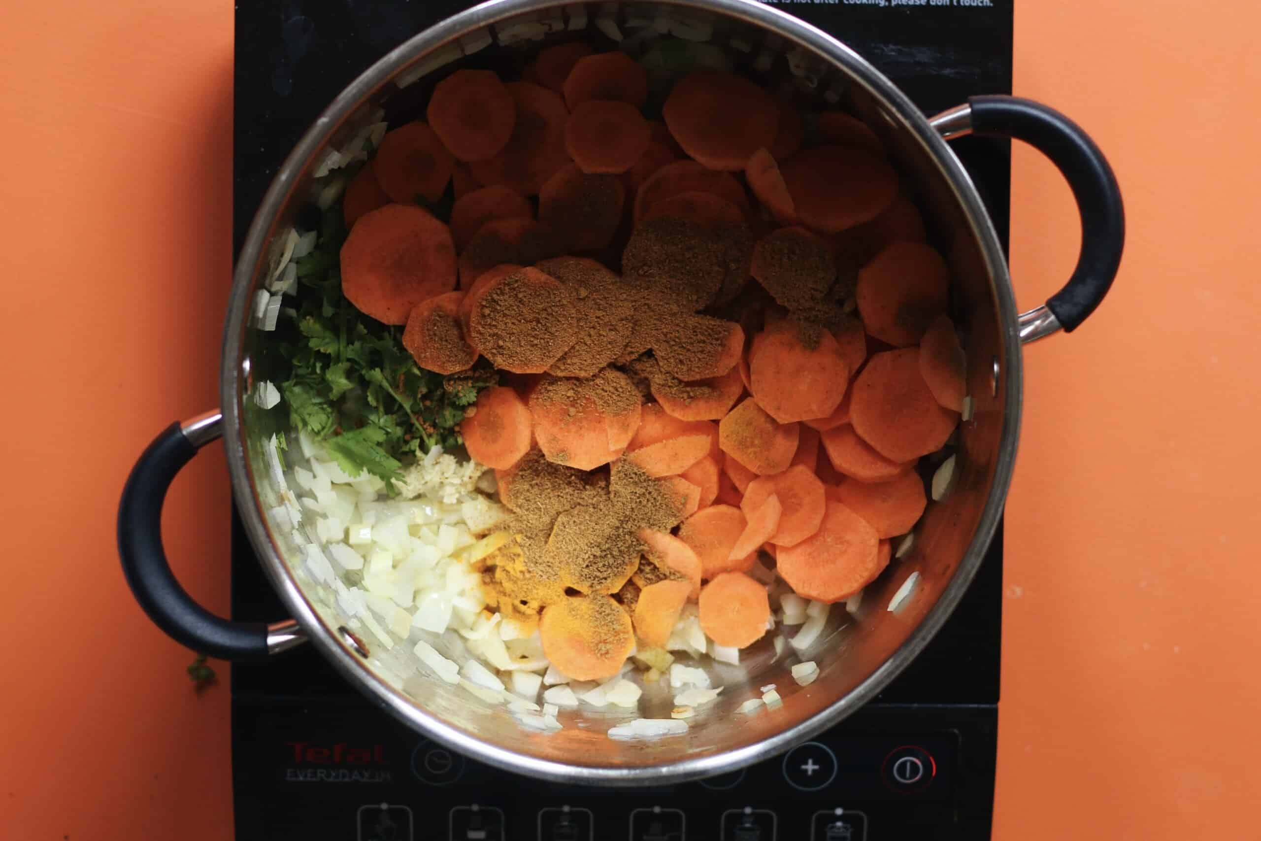 Carrots, spices, onions, lentils and coriander in large saucepan on a stove on an orange background.