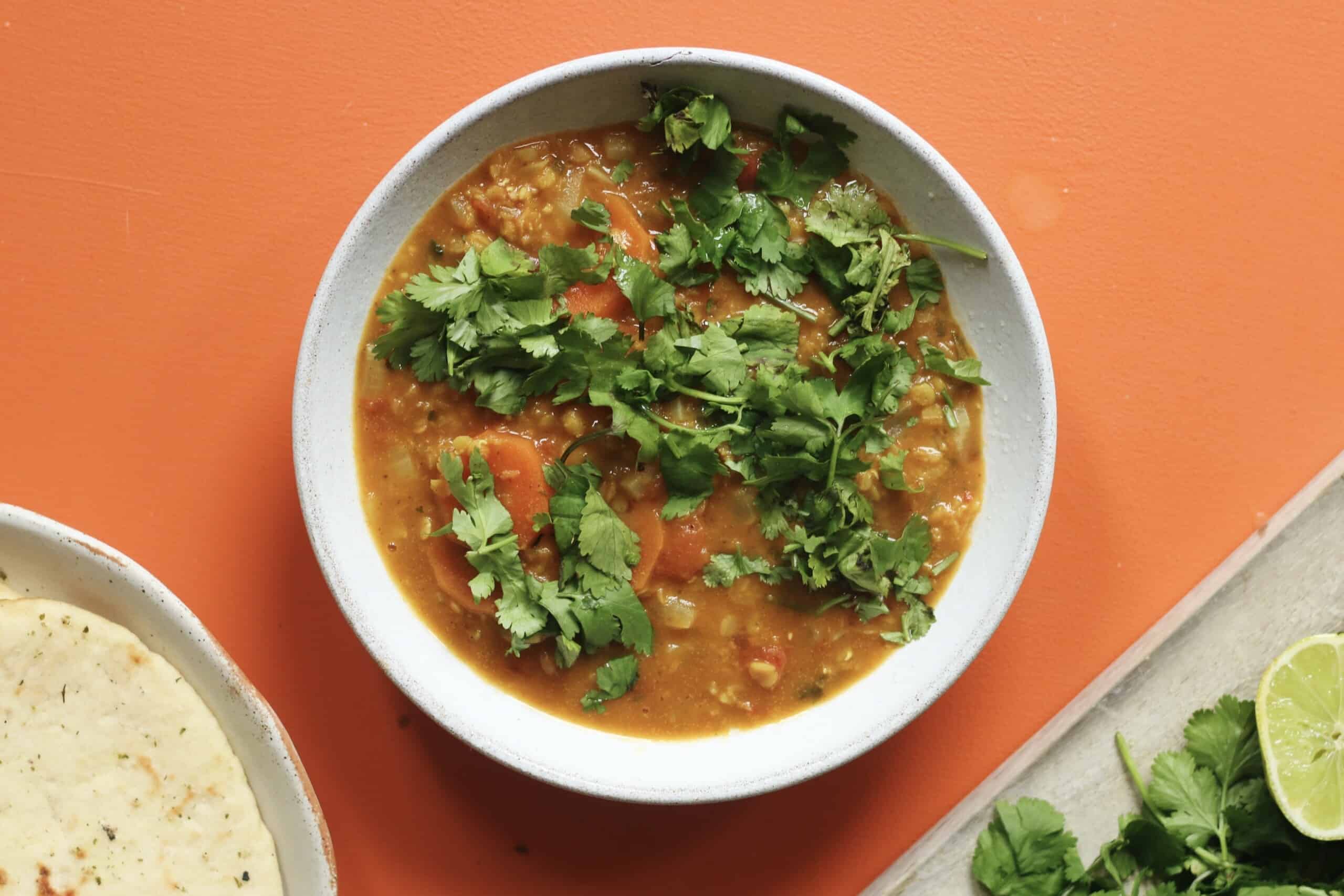 A bowl of red lentil dahl topped with lots of coriander with a separate bowl of naan and coriander and lime.
