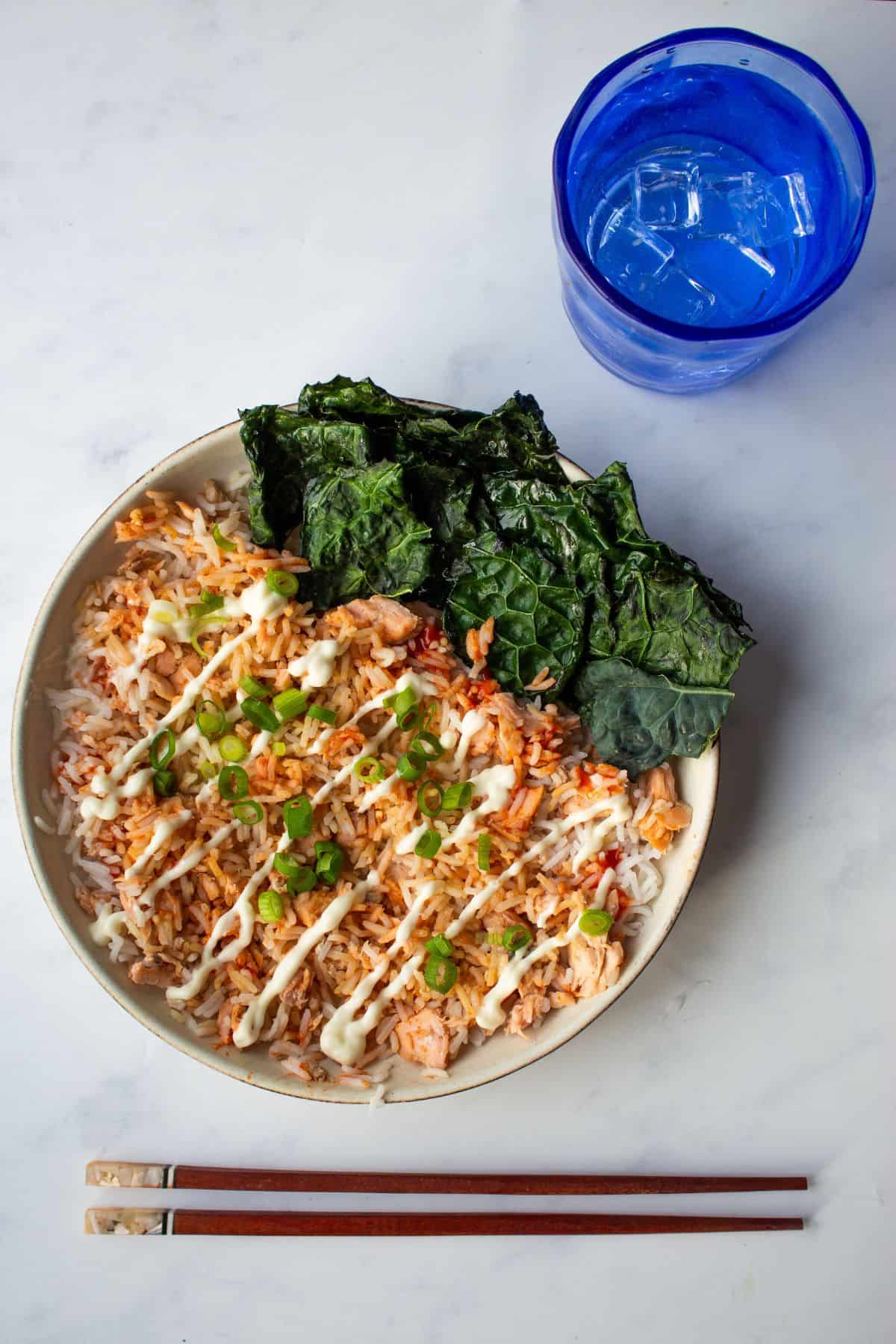 Salmon rice bowl served with kale with a blue glass with water and chopsticks next to the bowl.