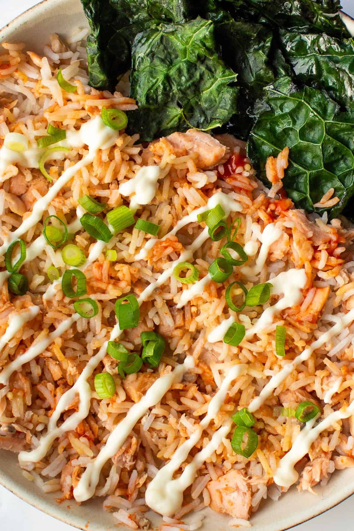 Close up of a bowl of rice with salmon served with a drizzle of mayonnaise, sliced spring onions, Sriracha and kale.