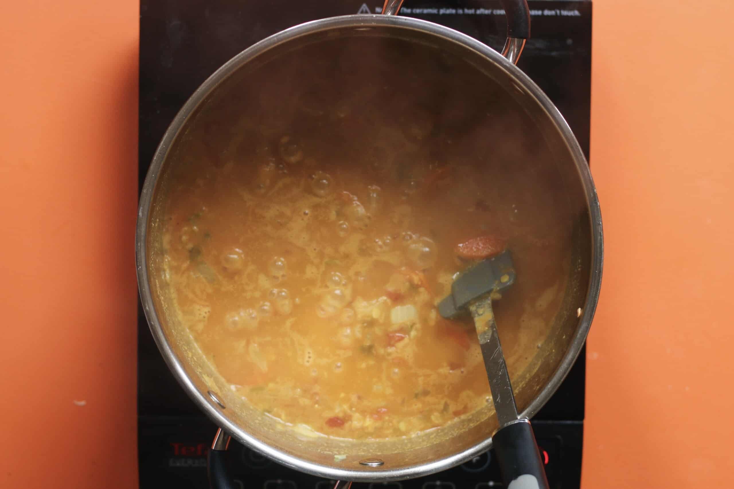 Red lentil dahl cooking a pan and stirred with a spatula on a stove on an orange background.